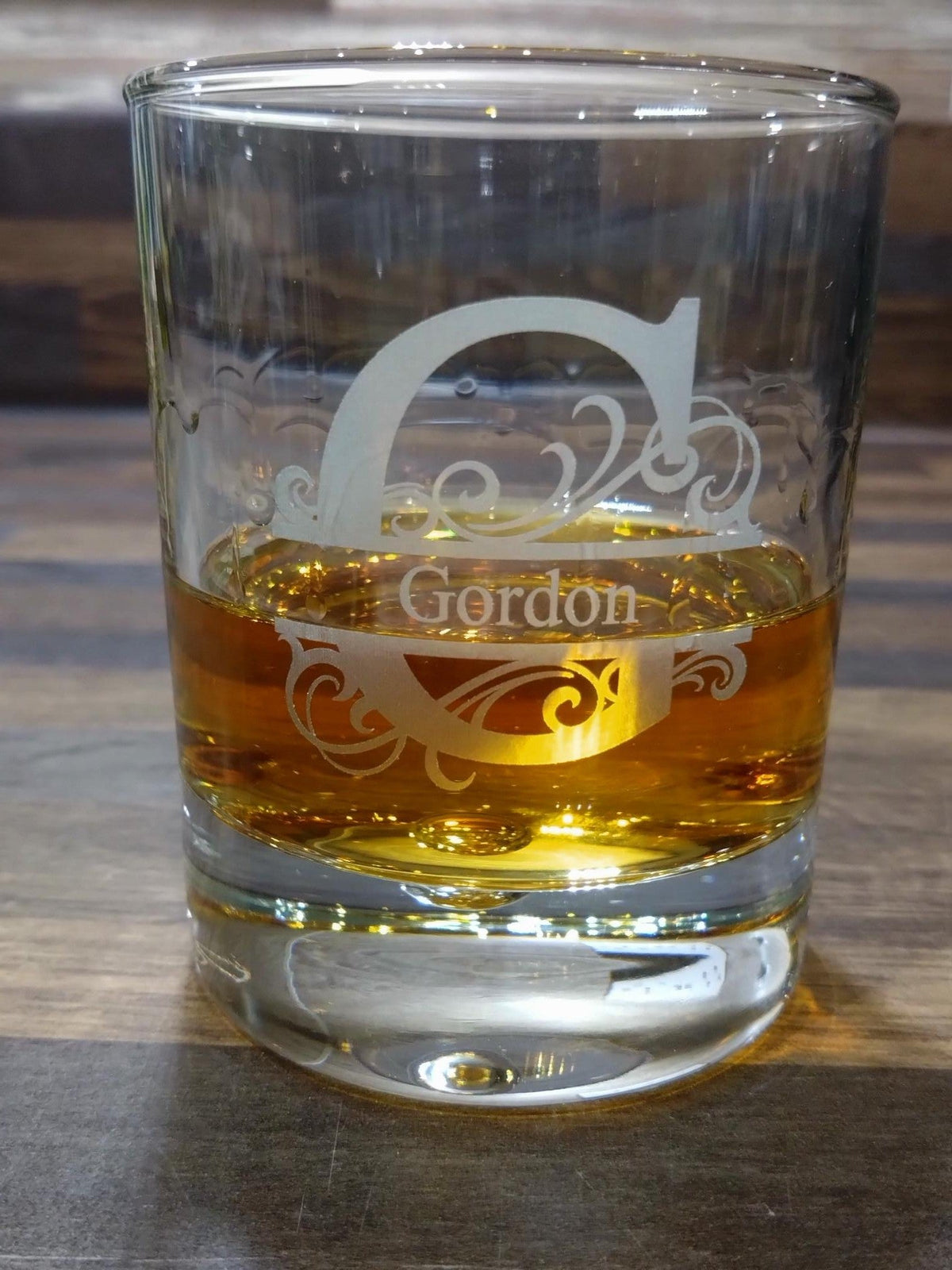 Glass tumbler with 'Gordon' engraving filled with a yellow liquid on a wooden surface.
